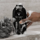 Black bottle of Mad Rabbit Refresh Body Wash with a hand holding it, surrounded by bath products on a wooden surface.