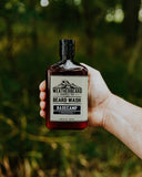 A hand holding a bottle of Weatherbeard Supply Co. Beard Wash Basecamp in front of a blurred green outdoor background.