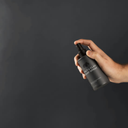 A hand holding a matte black spray bottle labeled 'SALT & CLAY SPRAY' against a dark gray background.
