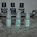 Three small perfume bottles with labels on a textured surface, with more bottles in the background.