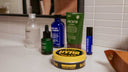 A collection of personal care and grooming products arranged on a bathroom counter, including a green Jackfir body wash tube, a green dropper bottle, a Blu Atlas face wash bottle, a Comune frosted bottle, a green bottle, and a yellow tin of Byrd Slick pomade.