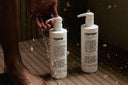 Two bottles of shampoo and conditioner being held over a shower with water splashing.