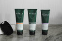Three Heath hair and body wash bottles on a marble surface with a textured wall background.