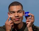Man applying cream to his face with a jar of skincare product in front of a blue background